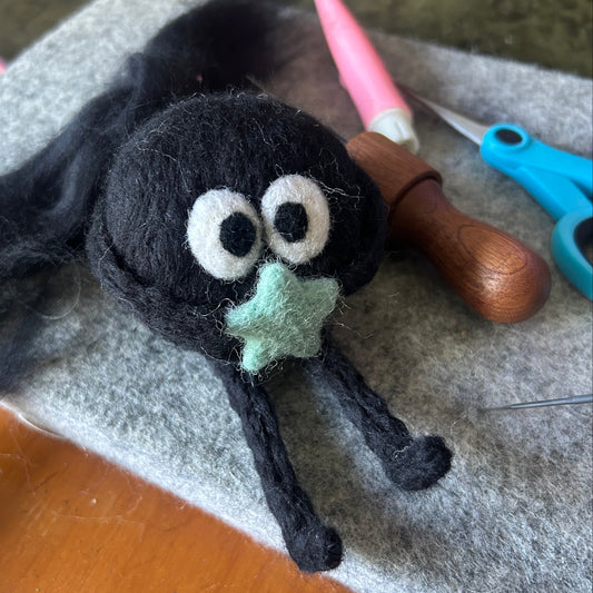 Handmade soot spirit inspired felt cat toy made with black wool, surrounded by hand felting tools